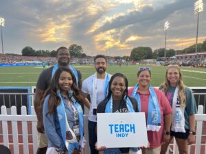 A diverse group of six smiling individuals standing together at an outdoor sports stadium, holding a sign that reads 'Teach Indy.'