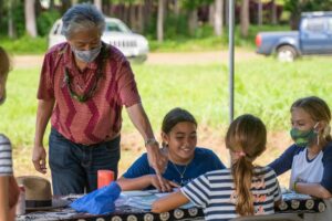 An older adult wearing a red shirt and a face mask interacts with three young children seated at a table under a canopy. The children are engaged in what appears to be an educational activity or craft project. They are surrounded by lush greenery in an outdoor setting, emphasizing a community or educational event.